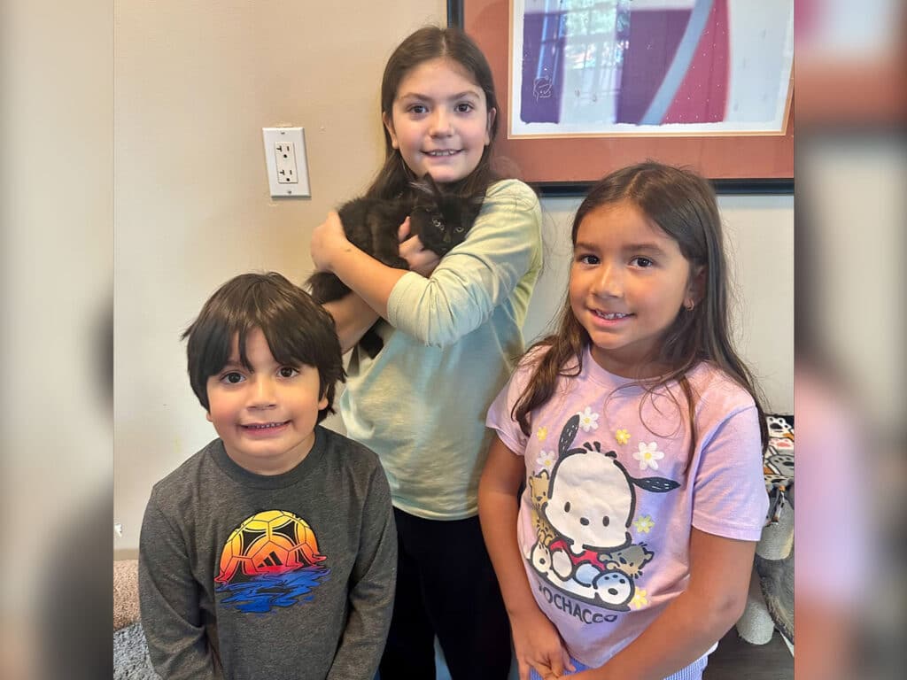 Three children are posing together in a well-lit room. The oldest girl holds a black kitten, while a boy and a younger girl stand next to her, smiling. The boy wears a gray shirt with a colorful graphic, and the girl has on a pink shirt with animal designs.