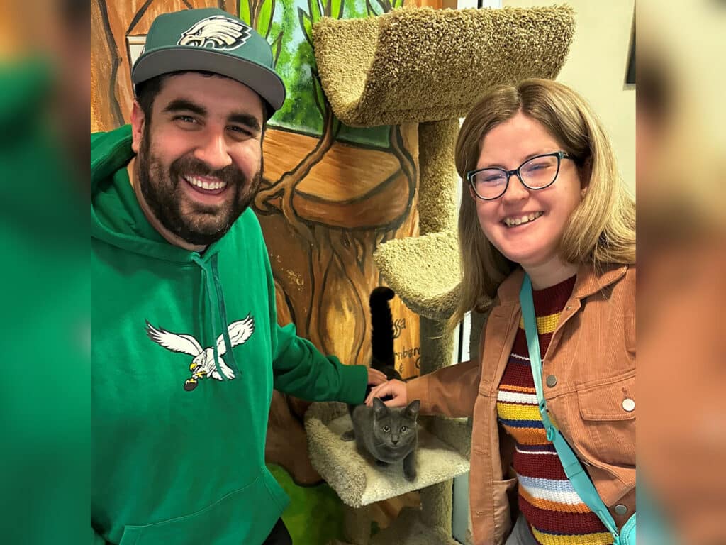 A man in a green hoodie and a woman wearing glasses and a brown jacket smile while standing next to a cat tree. A gray cat sits on a lower level of the tree, while the man gently pets it. The background features a colorful mural.