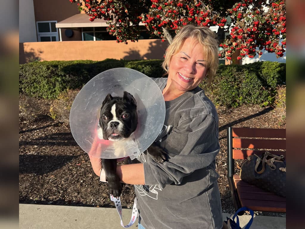 A woman with short blonde hair is smiling while holding a black and white dog that is wearing an Elizabethan collar. The background features green bushes and a building, indicating an outdoor setting.