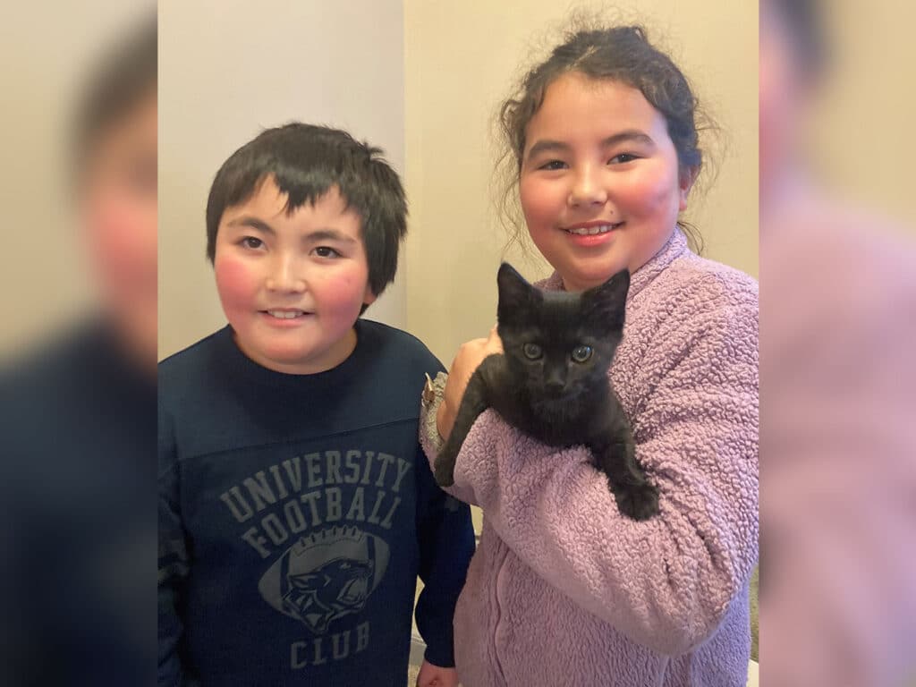 A young boy wearing a navy blue sweatshirt and a young girl in a light purple fleece hold a black kitten between them. They are smiling and standing in a well-lit room with neutral-colored walls.