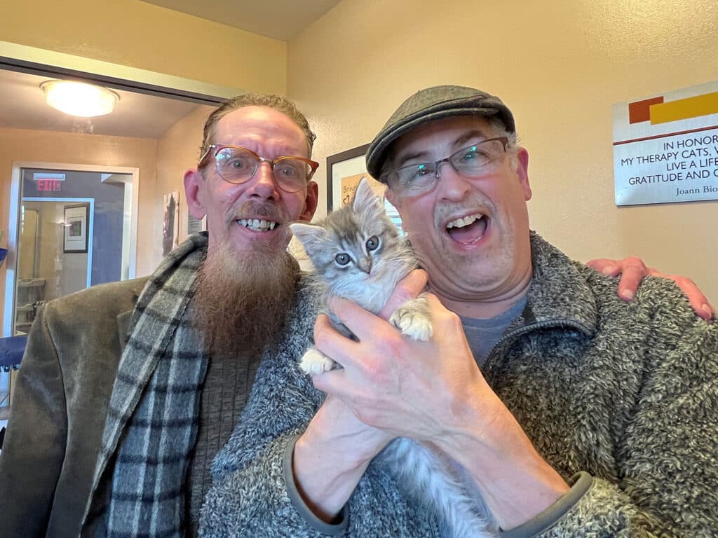 Two men smiling and holding a small gray and white kitten in a cozy indoor setting. One man has a beard and glasses, while the other wears a cap. A wall with a framed message is visible in the background.
