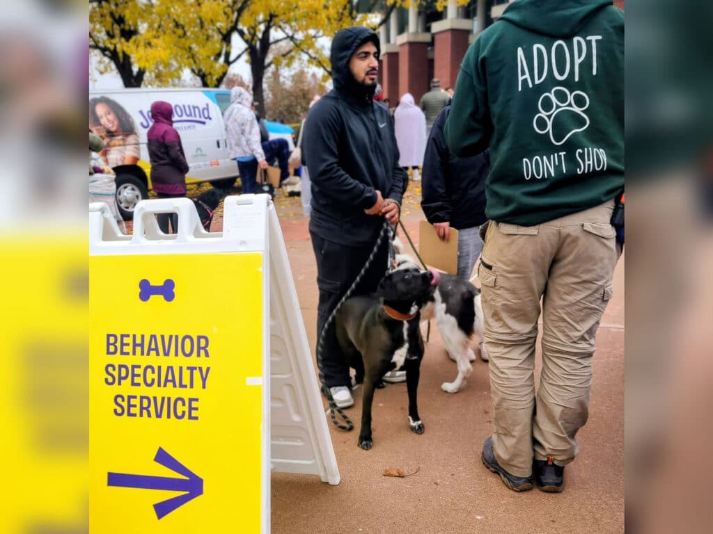 A man holding a dog's leash stands next to another person in a hooded sweatshirt, at an outdoor event. A sign with the text 'Behavior Specialty Service' points left, while an 'Adopt Don't Shop' graphic is visible on the sweatshirt.