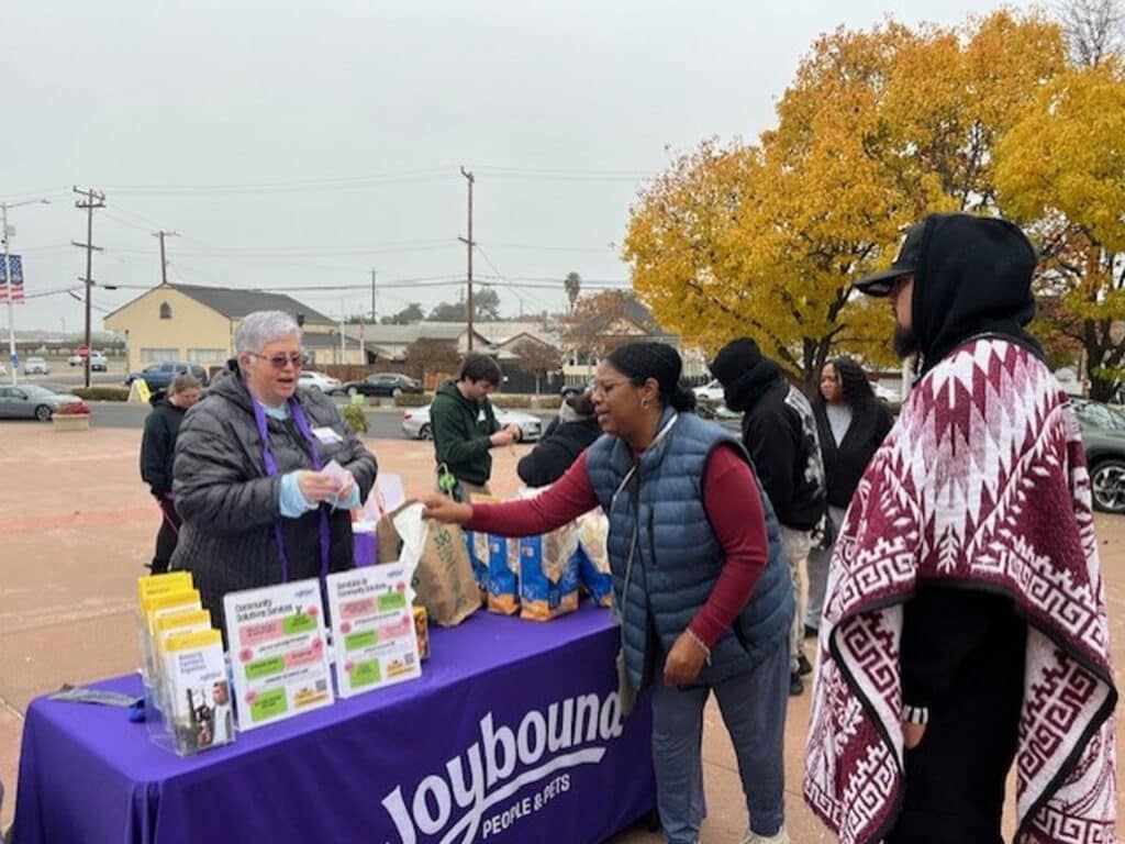 A woman is interacting with another woman at a table displaying vaccine information and materials. Several individuals are seen in the background, and autumn trees with yellow leaves are visible. The setting appears to be an outdoor community event.