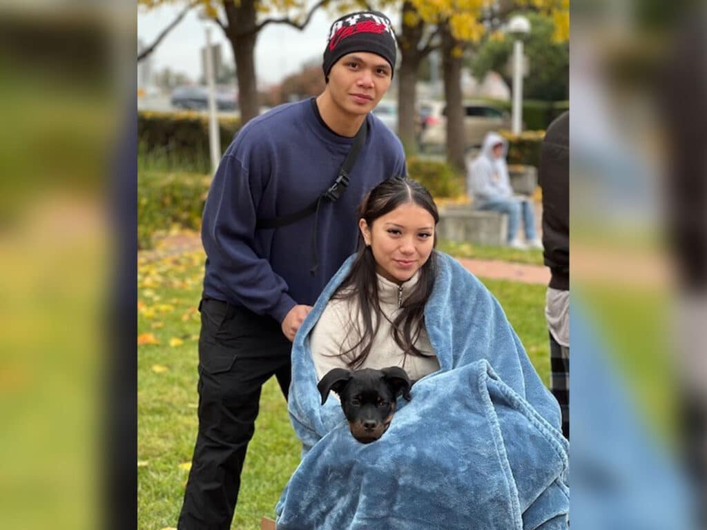 A young man in a dark sweater and beanie stands behind a young woman wrapped in a blue blanket, who is holding a small black dog. The scene is set in a park with autumn trees in the background.