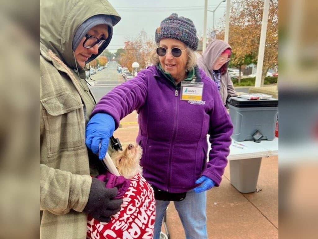 A woman wearing a purple jacket and gloves administers a vaccination to a small dog held by a person in a green jacket. Another person is in the background, and autumn foliage is visible in the surrounding area.