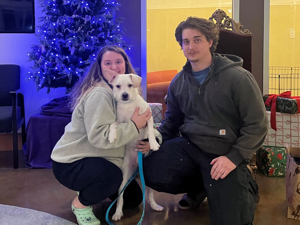 A woman and a man kneel beside a small, light-colored dog, smiling. The woman wears a gray sweater and black pants, while the man has on a dark hoodie and black pants. A decorated Christmas tree is visible in the background.