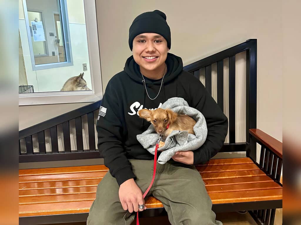 A young man wearing a black beanie and hoodie is seated on a wooden bench, holding a small dog wrapped in a gray blanket. The background includes a pet adoption area with a glimpse of another animal.