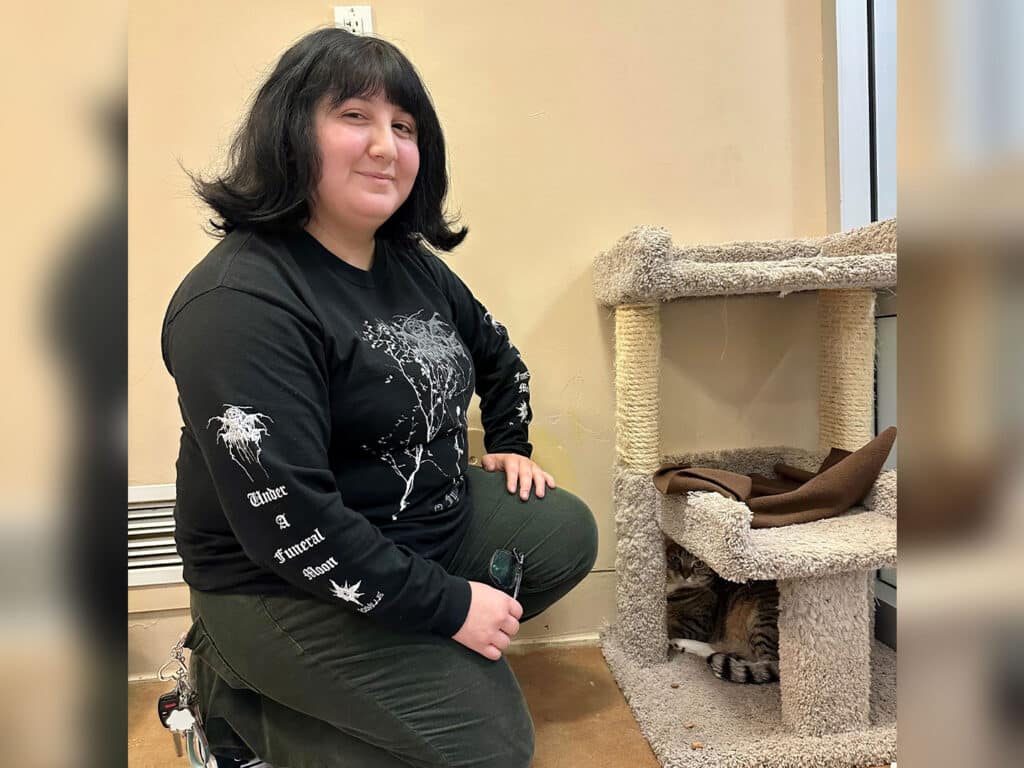 A woman with long dark hair is kneeling beside a cat tree in a cozy indoor space. She wears a black long-sleeve shirt with white designs. A brown blanket lies on a ledge of the cat tree, and a tabby cat is partially visible inside.