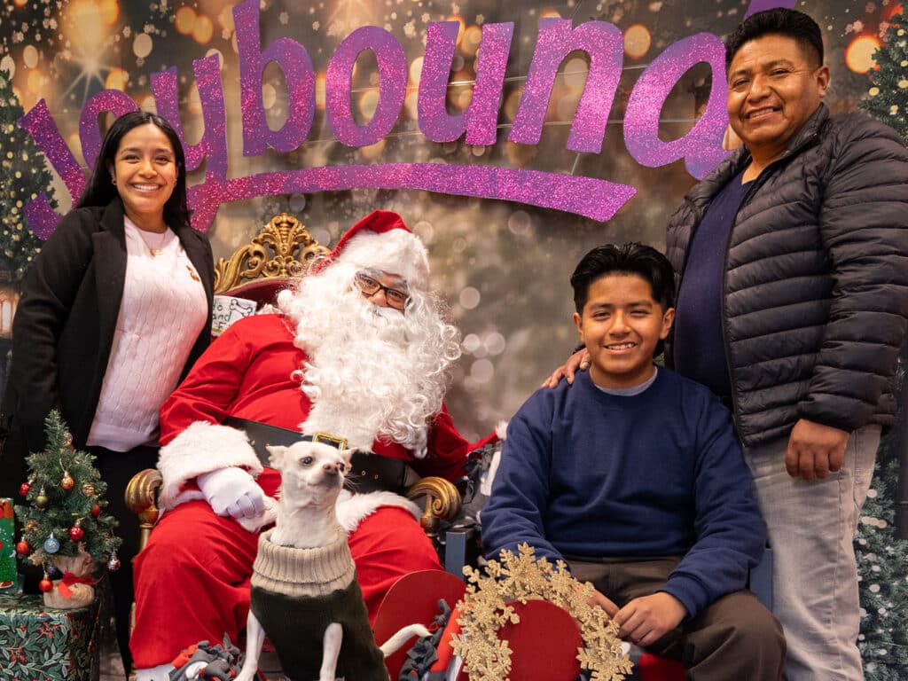 A family poses with Santa Claus in a holiday-themed setting. Santa is seated in a chair, wearing a traditional red suit and white beard. To his left, a woman and a boy stand, and to his right, a man stands next to a small dog. The background features festive decorations.