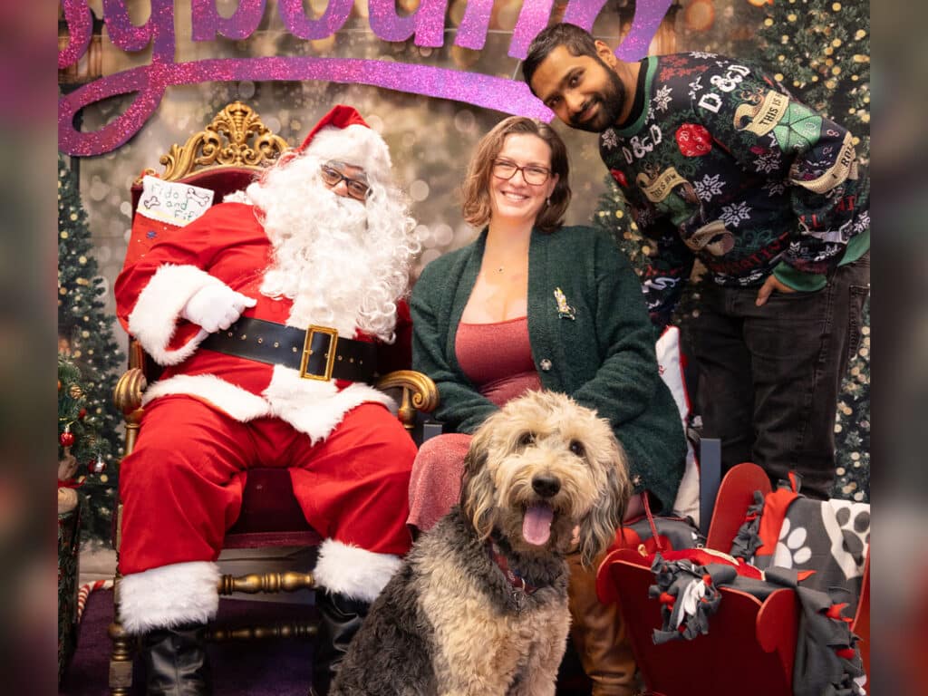A woman in a green cardigan and red top sits next to a large dog in front of Santa Claus, who wears a traditional red suit. A man in a festive sweater stands beside them, smiling. The background is decorated with Christmas-themed elements.
