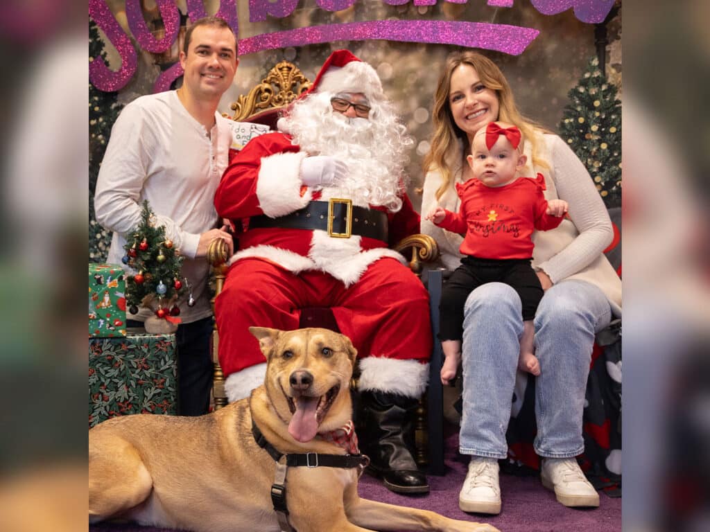 A family poses with Santa Claus at a holiday-themed photoshoot. The group includes two adults, a baby in a red outfit, and a dog. The backdrop features festive decorations, including gifts and a Christmas tree.
