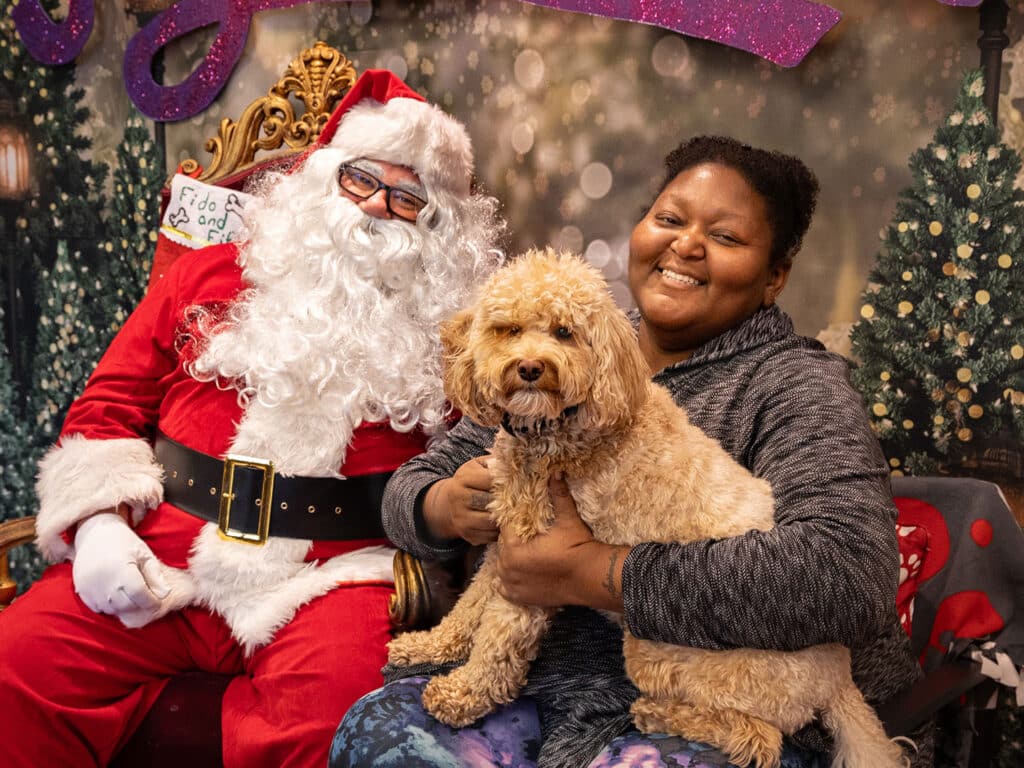 A woman sitting on a couch, smiling with a small light-brown dog in her lap. Next to her is a man dressed as Santa Claus, featuring a white beard and red suit. The background includes green trees and holiday decorations.