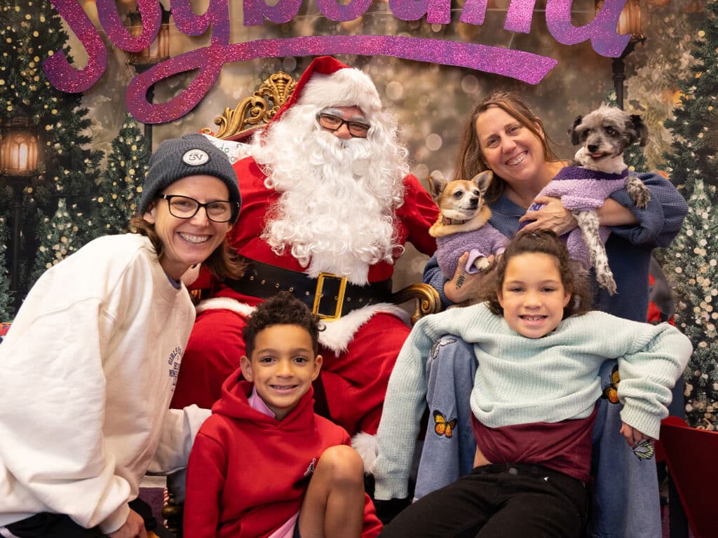 A group photo featuring two children, a woman, and Santa Claus, with two small dogs. The children sit in front, smiling, while the woman holds one dog. The backdrop displays festive decorations with the word "Joy" prominently featured.