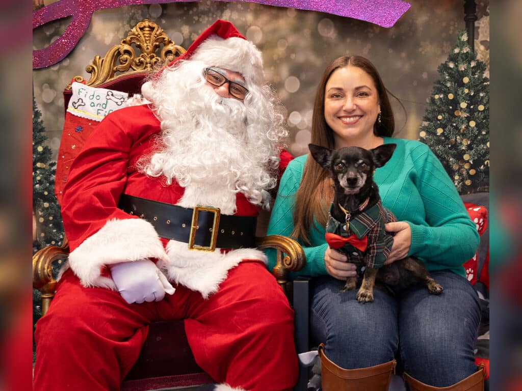 A woman with long brown hair wearing a teal sweater sits on Santa Claus's lap, holding a small dog dressed in a plaid outfit. Santa, in a classic red suit with a white beard, smiles next to Christmas decorations in the background.
