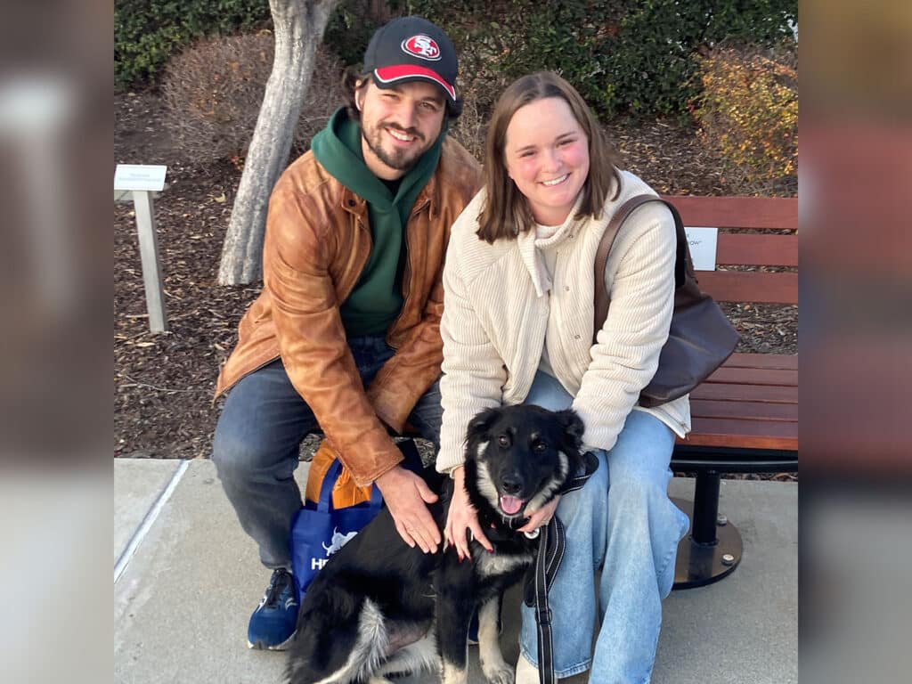A man and a woman sit on a bench outdoors, with a medium-sized black and white dog in front of them. The man wears a brown leather jacket and a cap, while the woman wears a cream-colored coat. They are smiling and appear to be in a park-like setting.