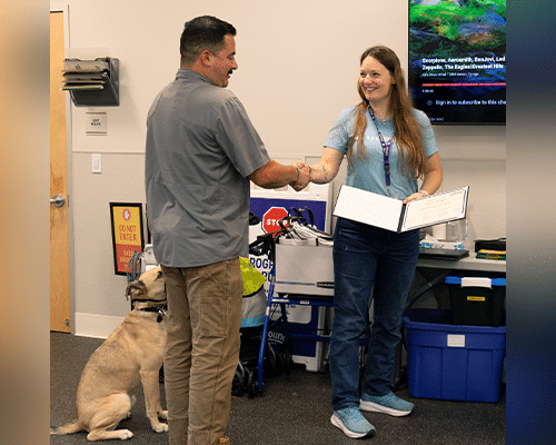 A man in a gray shirt shakes hands with a woman in a blue shirt holding a certificate in a room with various items in the background. A dog sits nearby on the floor. The setting appears to be a ceremony or celebration.