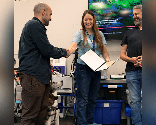 A man shakes hands with a woman in a blue shirt who is handing him a certificate. To the right, another man smiles. A service dog stands nearby in front of covered storage boxes, with a projector screen visible in the background.