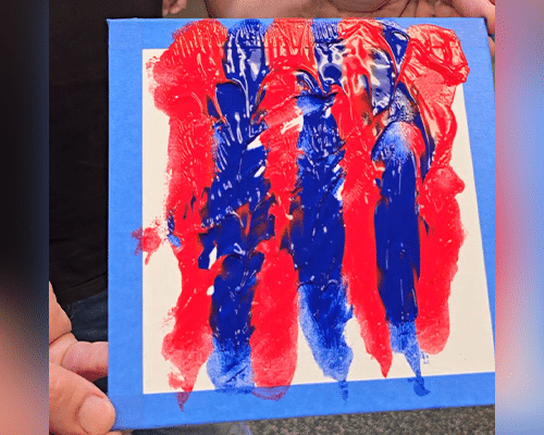 A hand holding a textured painting on canvas, featuring bold red and blue brush strokes on a white background. The painting is bordered by blue tape, indicating it may be a fresh artwork or part of an art project.
