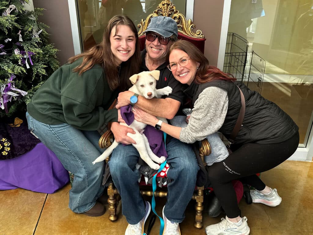 A man sitting on a golden chair holds a small white puppy, surrounded by two women. One woman is smiling and sitting on his left, while the other woman, wearing a black vest, hugs him from the right. A decorated Christmas tree is visible in the background.