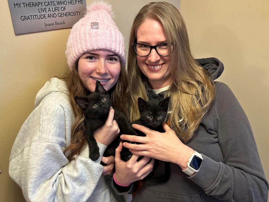 Two women are holding black kittens in their arms, standing in a cozy indoor setting. One woman wears a pink knit beanie and a gray sweatshirt, while the other has long hair and glasses, dressed in a darker top. A framed quote about therapy cats is visible on the wall behind them.