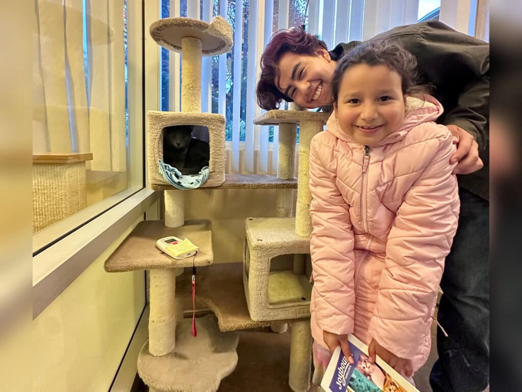 A young girl in a pink jacket smiles while standing next to a cat tree. Behind her, a boy with dark hair leans in, both posing for the photo. A black cat is visible inside a cubby on the tree. The setting is bright with large windows.