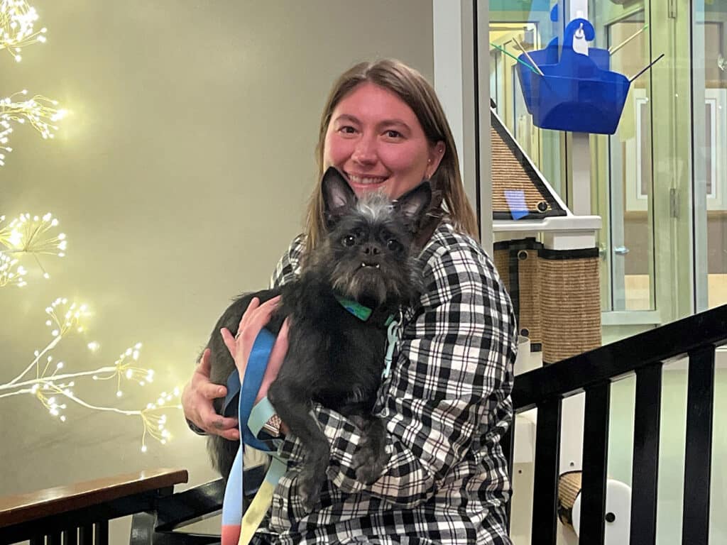 A woman sitting on a wooden chair holds a small black dog with a gray face. She has long brown hair and is wearing a black and white checkered shirt. Soft, decorative lights are visible in the background, creating a warm atmosphere.