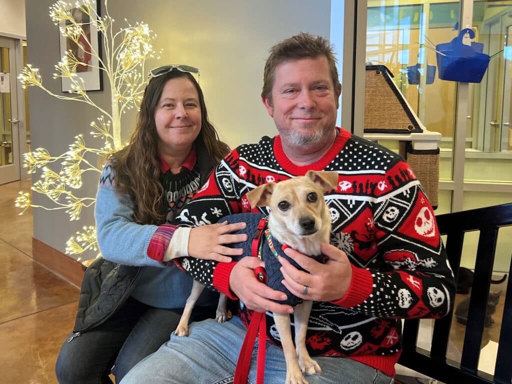 A woman and a man sitting on a bench in a warm indoor setting, both wearing festive sweaters. The woman has long dark hair and a gray vest, while the man has short light brown hair. They are holding a small dog on a leash.