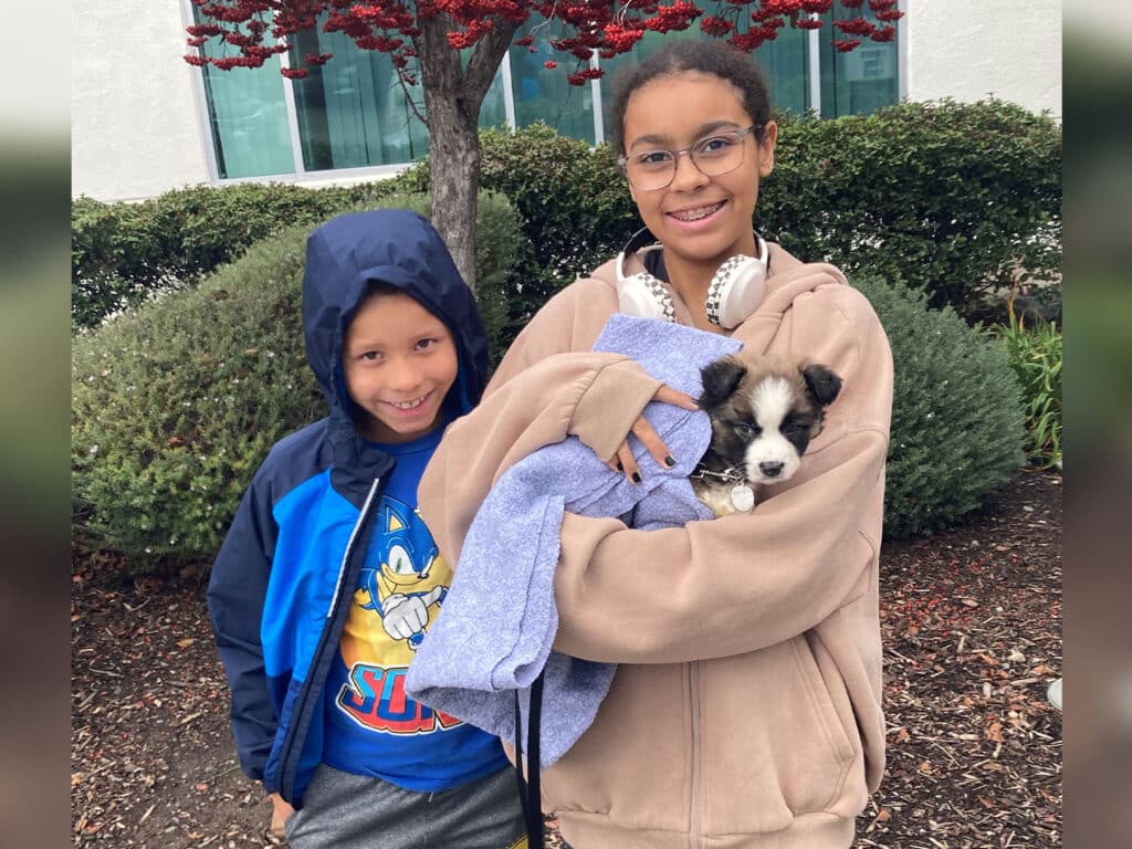 A girl holding a small puppy wrapped in a blue blanket stands beside a smiling boy outdoors, with greenery and a building in the background. The girl wears a beige hoodie and headphones, while the boy is dressed in a blue shirt.