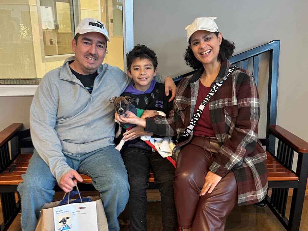 A family of three sits on a wooden bench inside a building. A man and a woman smile at the camera, holding a small puppy in a blanket. A young boy beside them grins while reaching for the puppy. They are casually dressed and the setting is bright.