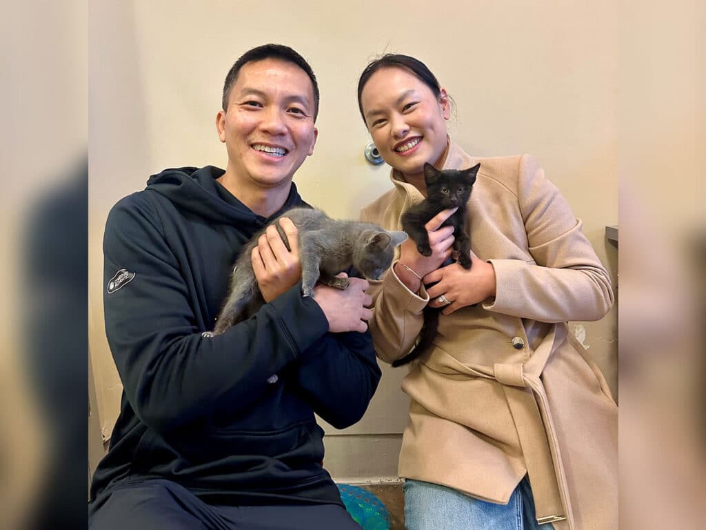 A man and a woman smiling while holding two kittens. The man has short hair, wearing a black hoodie, and the woman is in a beige coat with long hair. They are in front of a light-colored wall, with a playful atmosphere.