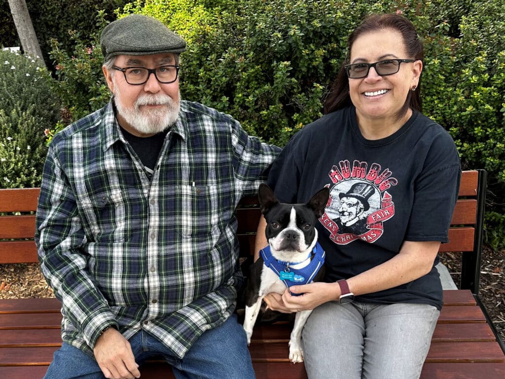 A man and a woman sitting on a park bench, smiling while holding a small black and white dog. The man wears a plaid shirt and a flat cap, while the woman wears a graphic t-shirt and glasses. Green plants are visible in the background.