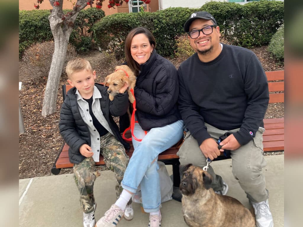 A woman, a boy, and a man sit on a bench outdoors, posing with a small dog held by the woman. The boy wears a gray and black jacket with camo pants, while the man wears a black sweater. A larger dog is beside the man on the ground.