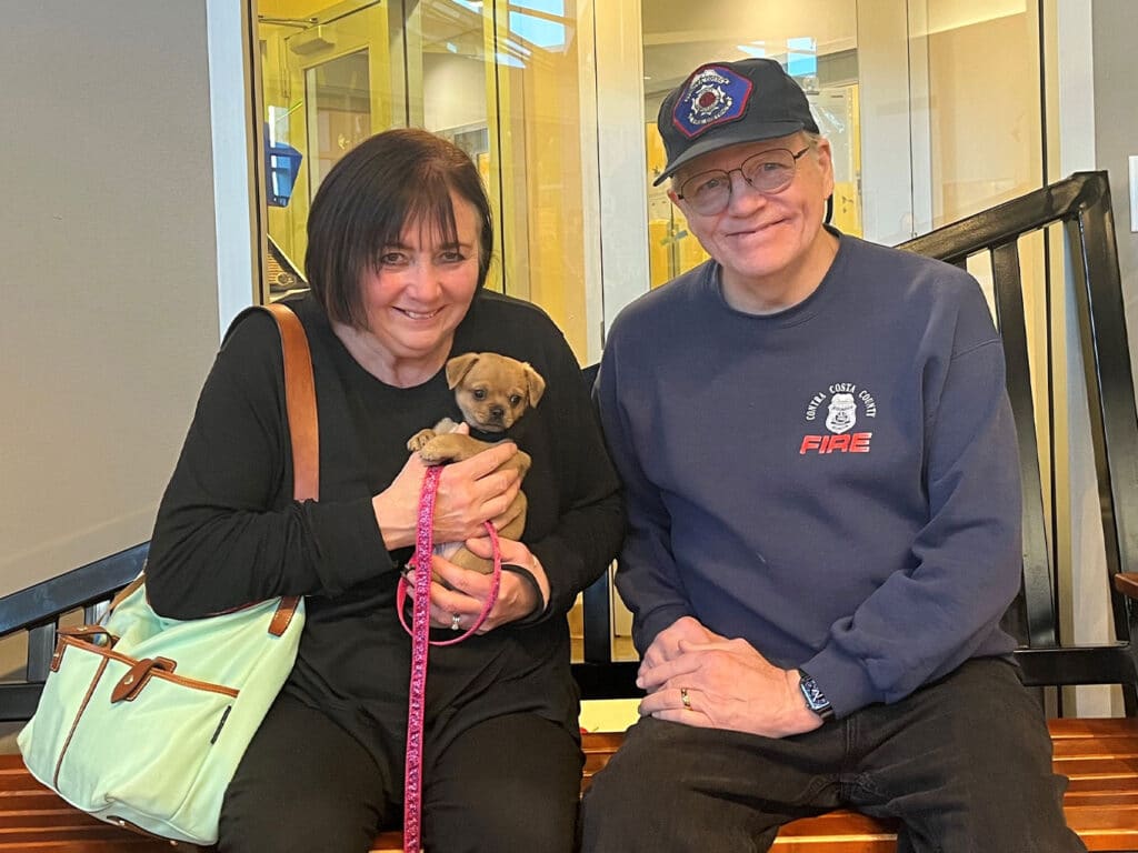 A woman and a man sitting on a bench inside a building, holding a small puppy. The woman is wearing a black shirt and holding the puppy in her arms, while the man is wearing glasses and a navy sweater. A mint green bag is beside them.