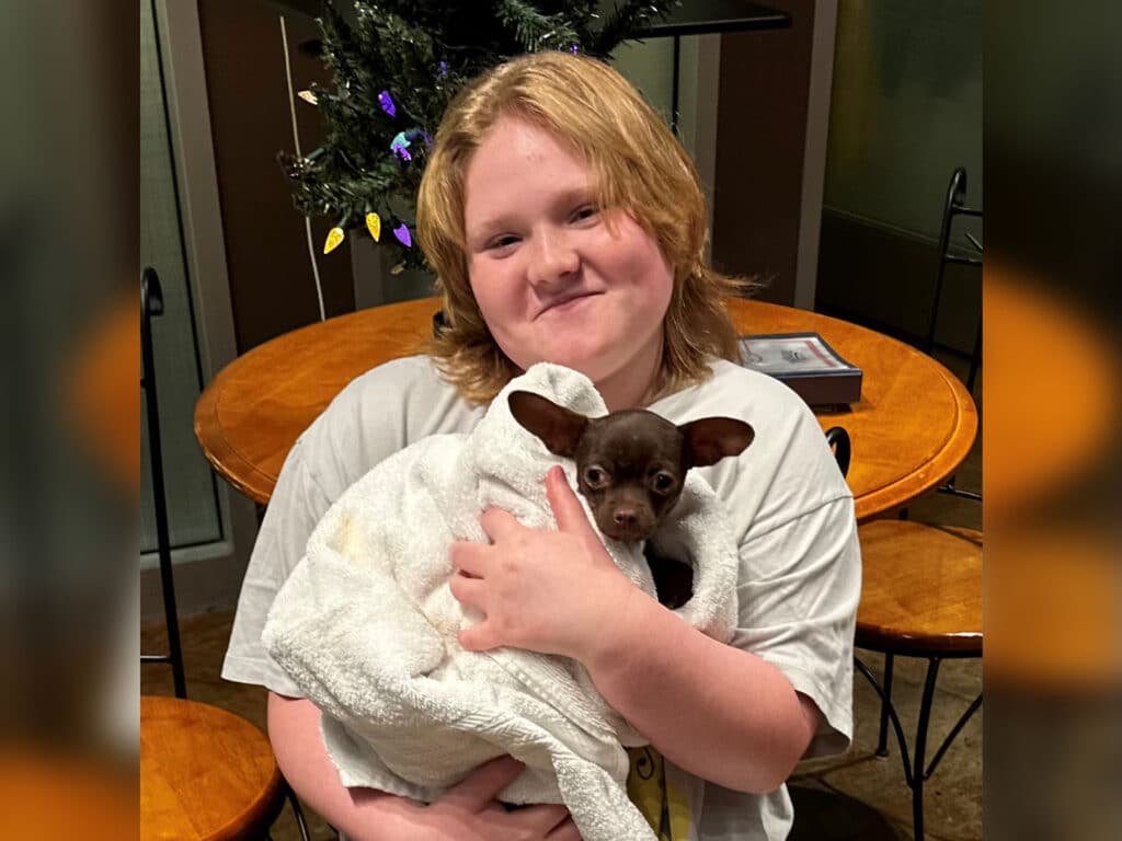 A young person with shoulder-length blonde hair smiles while holding a small, brown dog wrapped in a white towel. They are indoors, with a wooden table and Christmas decorations in the background, creating a cozy setting.