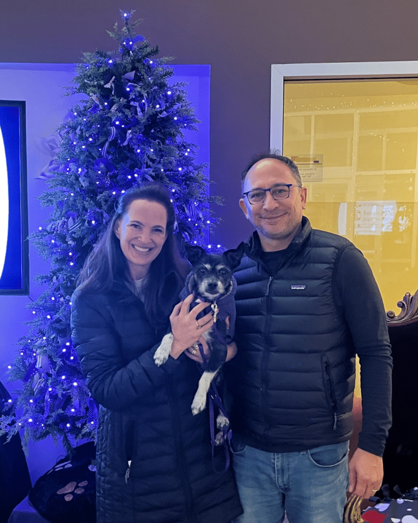A woman and a man stand together in front of a decorated Christmas tree with blue lights. The woman holds a small dog while both smile at the camera. The setting features a festive atmosphere with holiday decorations visible.