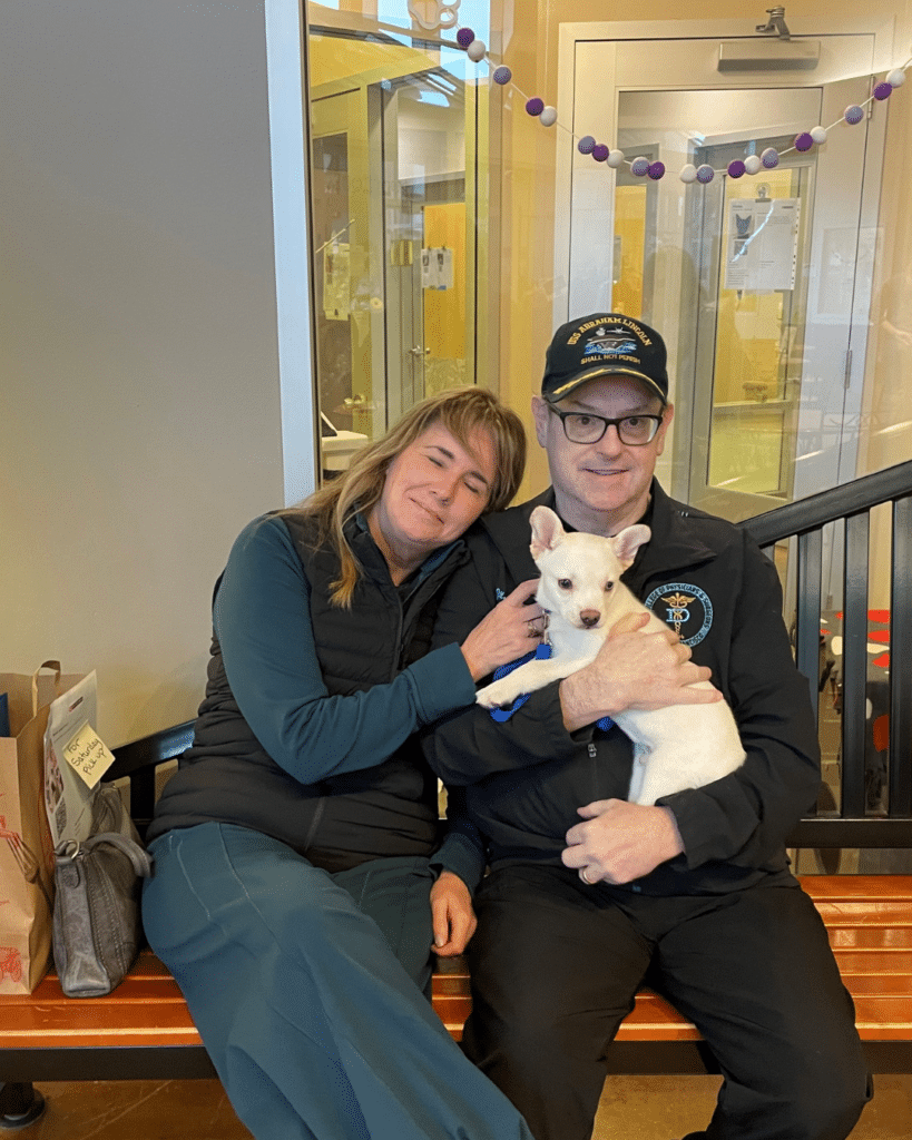 A woman and a man sit together on a bench in a lobby, each holding a small white dog. The woman has long hair and is wearing a dark vest and green pants. The man, wearing a black jacket and a cap, is smiling at the camera.