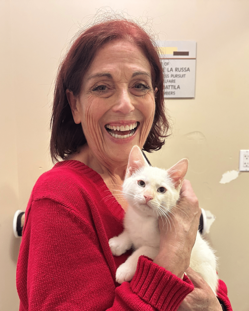 A woman with shoulder-length brown hair, wearing a red sweater, smiles widely while holding a white kitten close to her face. The background features a plain wall with a visible sign.