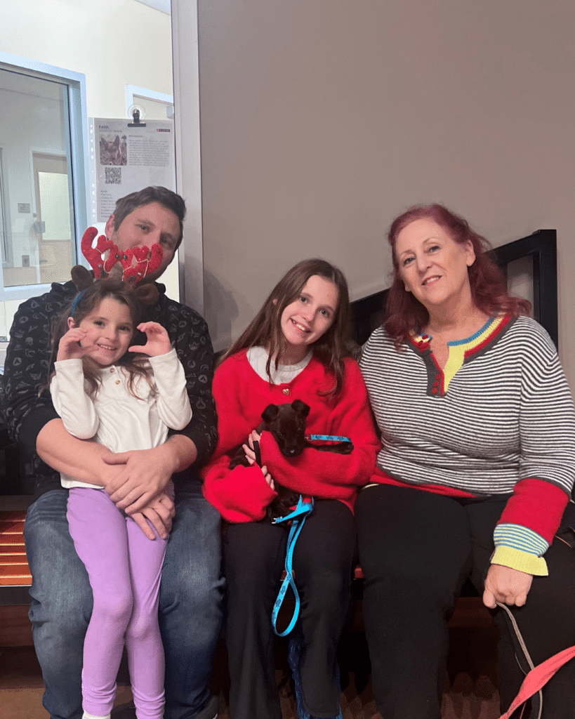 A family of four sitting together in a waiting area. The father has reindeer antlers, the younger girl is wearing a white sweater, the older girl is dressed in a red sweater holding a small dog, and the woman has red hair and a striped shirt.