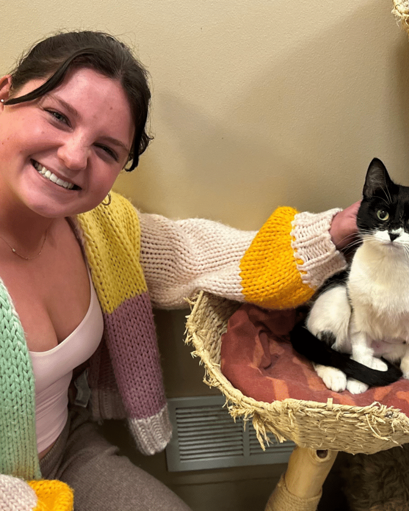A young woman with dark hair smiles while sitting next to a black and white cat on a cat tree. The woman wears a pastel-colored knitted sweater and leans slightly towards the cat, who is sitting calmly in a basket.