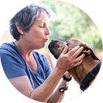 A volunteer interacting with a brown dog, gently pressing her forehead against the dog's and smiling. The background features greenery, suggesting an outdoor setting, reflecting a moment of connection between the volunteer and the animal.