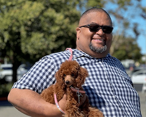 A man with sunglasses smiles while holding a small brown poodle on a leash. He wears a checked shirt and stands outdoors with trees and parked cars in the background.