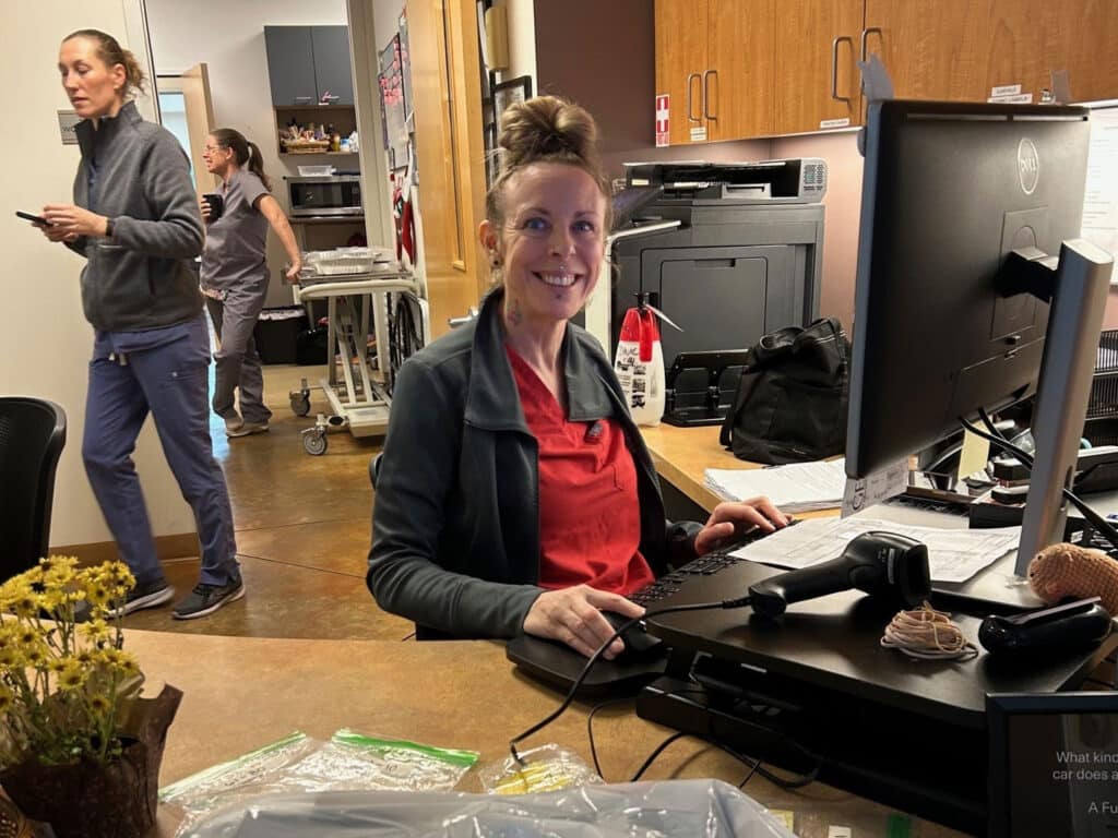 A woman with a bun hairstyle sits at a desk, smiling while using a computer. In the background, two other individuals are walking, one holding items. The workspace includes office equipment and a small bouquet on the desk.