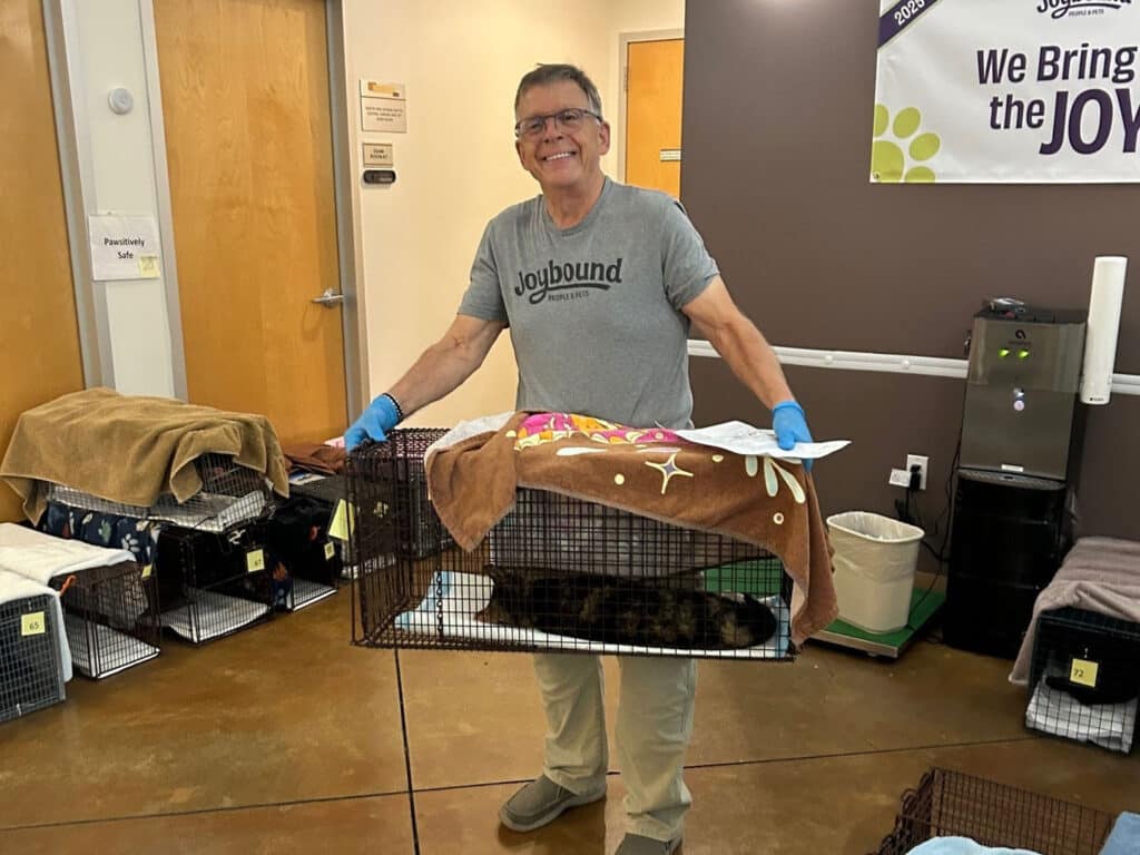 A smiling man holding a cat carrier wrapped with a blanket inside a room decorated with animal supplies. Various cat carriers are seen on the floor, and a banner that reads "We Bring the JOY" is visible in the background.