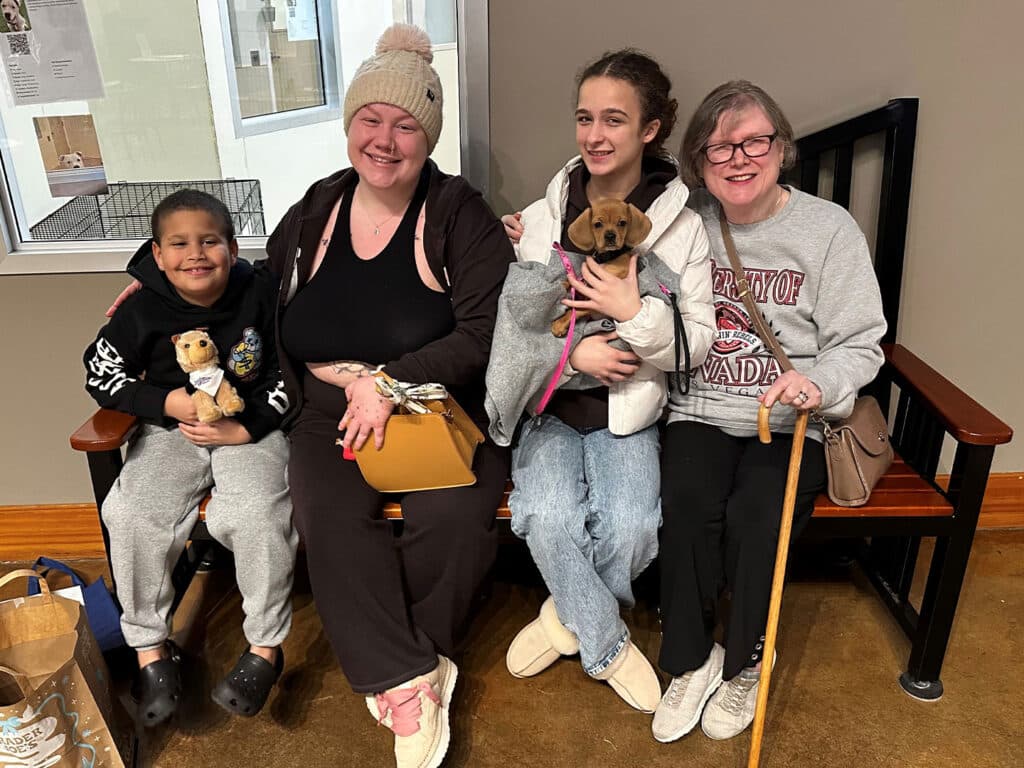 A family sits on a bench in a waiting area, smiling at the camera. A woman holds a puppy in her arms, while a girl next to her cradles it. A boy with a stuffed animal sits on the other side, beside a woman holding a bag and using a cane.
