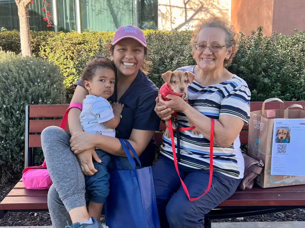 A woman holding a toddler on a bench, smiling. Next to them is an elderly woman with glasses holding a small puppy on her lap. They are surrounded by greenery and a notice board in the background, suggesting a community venue.