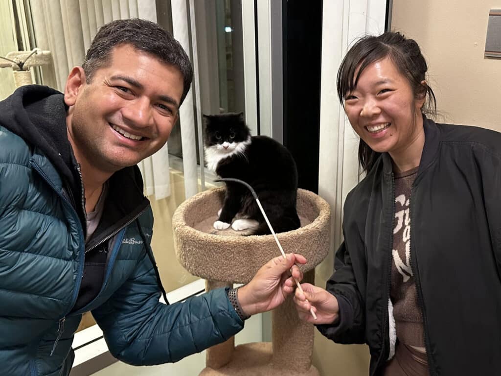 A man and a woman smile while posing for a photo in front of a cat sitting in a cat tree. The woman holds a cat toy on a stick, and the man stands beside her, both appearing cheerful in a well-lit space.