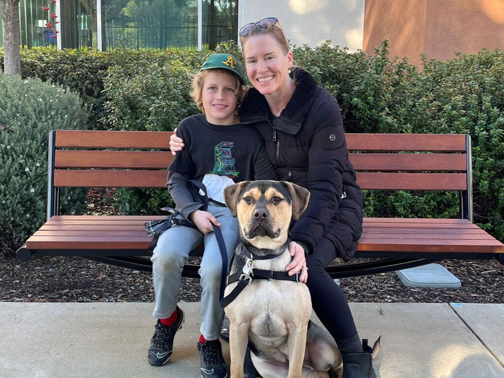 A woman and a boy sit on a wooden bench outdoors with a dog in front of them. The boy has his arm around the woman. The dog, a tan mixed breed, is on a leash. In the background, there are green shrubs and a building.