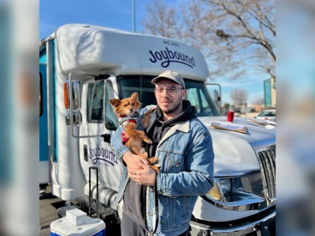 A man wearing glasses and a denim jacket holds a small brown dog in front of a mobile clinic vehicle marked "Joybound." The scene is set outdoors with trees visible in the background and clear blue sky.
