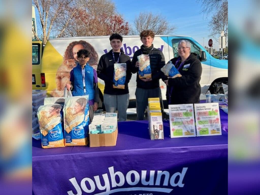 Four individuals stand behind a table displaying food packages and informational brochures. The backdrop features a vehicle with a colorful design. The group appears to be participating in a community event focused on food distribution.