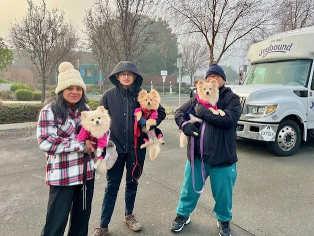 Three individuals standing outdoors in a chilly environment holding Pomeranian dogs. Two people wear winter clothing, including a beanie and a plaid jacket. A food truck is parked in the background among trees.